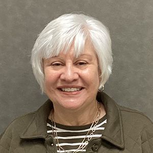 Woman with short white hair, smiling, wearing a striped shirt and quilted jacket.