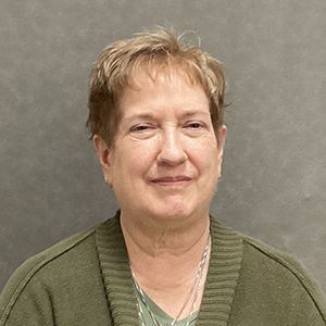 Woman with short blonde hair in a green sweater smiles slightly, in front of a gray backdrop.