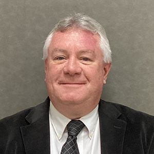 Man in suit and tie smiling, gray hair, indoors against a gray background.