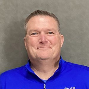 Man in blue collared shirt smiles, against a gray background.