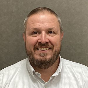 Man with beard smiles at the camera, wearing a light-colored collared shirt, gray background.