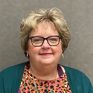 Woman wearing glasses, teal sweater, patterned blouse smiles at the camera.