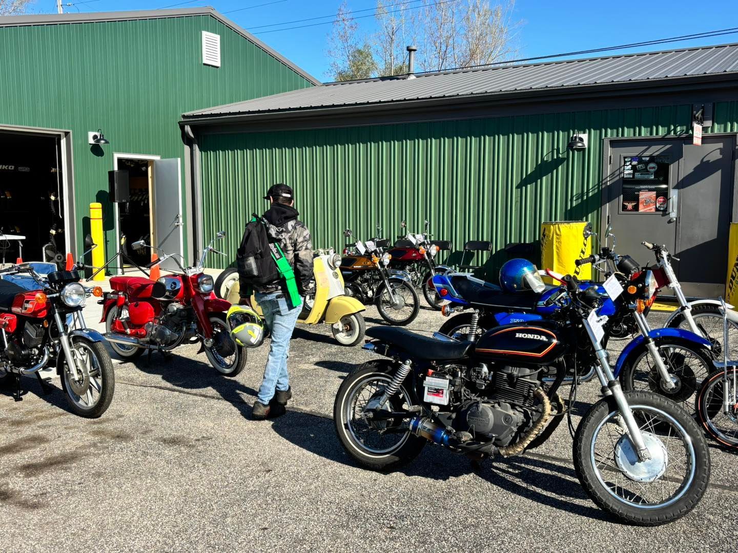 Person surrounded by several vintage motorcycles parked outside a green building on a sunny day.