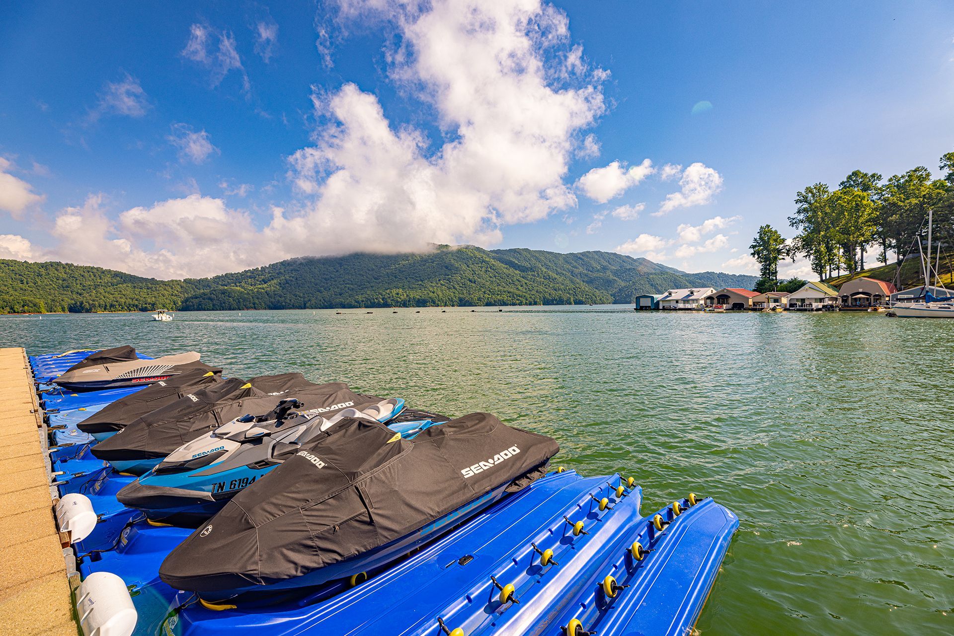 Covered jet skis on blue floating docks, with a lake and forested hills beyond
