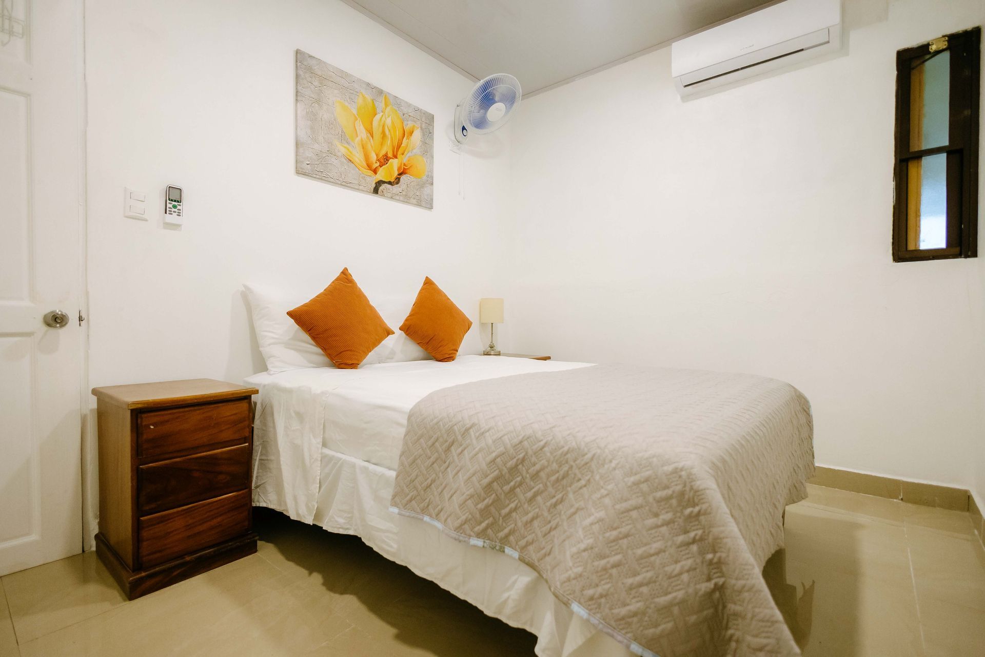 Habitación pequeña con cama blanca, almohadas naranjas, mesita de noche, cuadros en la pared y aire acondicionado.