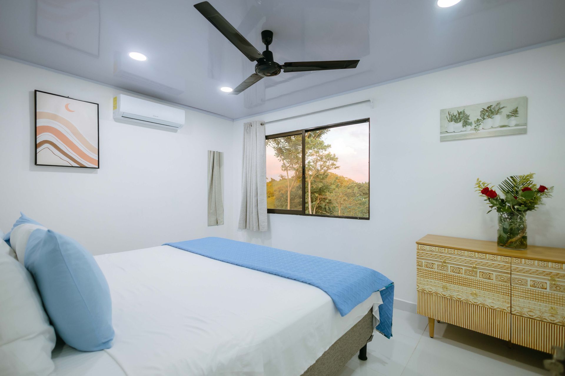 Habitación luminosa con cama blanca, detalles en azul, ventilador de techo, ventana y jarrón con flores sobre una mesa.