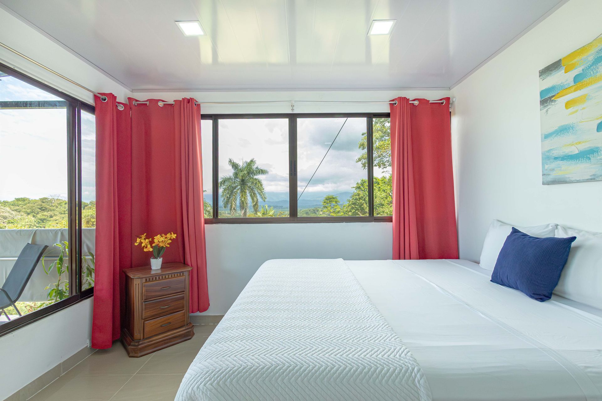 Habitación luminosa con cortinas rosas, cama blanca y vistas al océano a través de grandes ventanales.