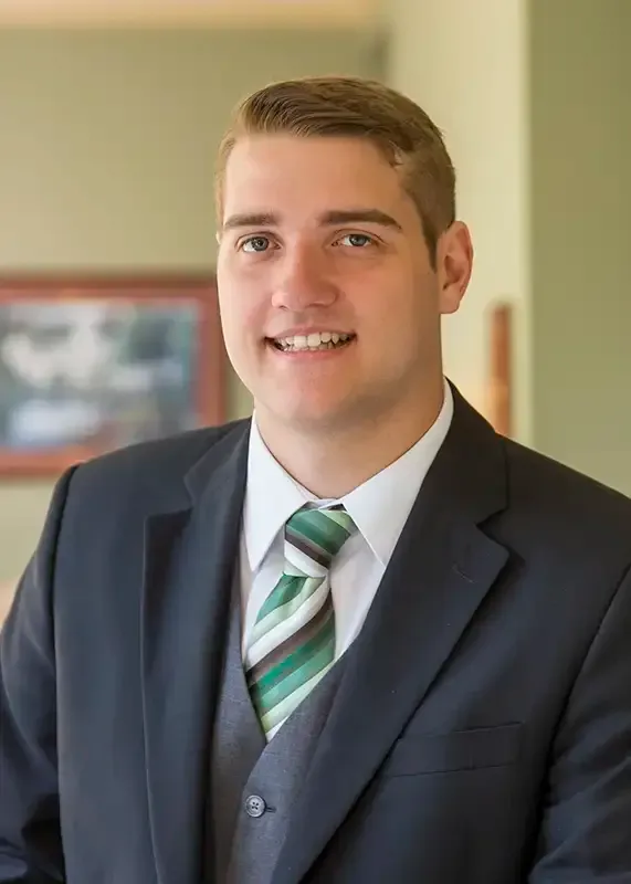 Man in a suit with a green striped tie smiles at the camera; indoor setting with a painting.