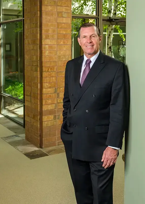 Man in a dark suit, leaning against a wall in an office setting, smiling.