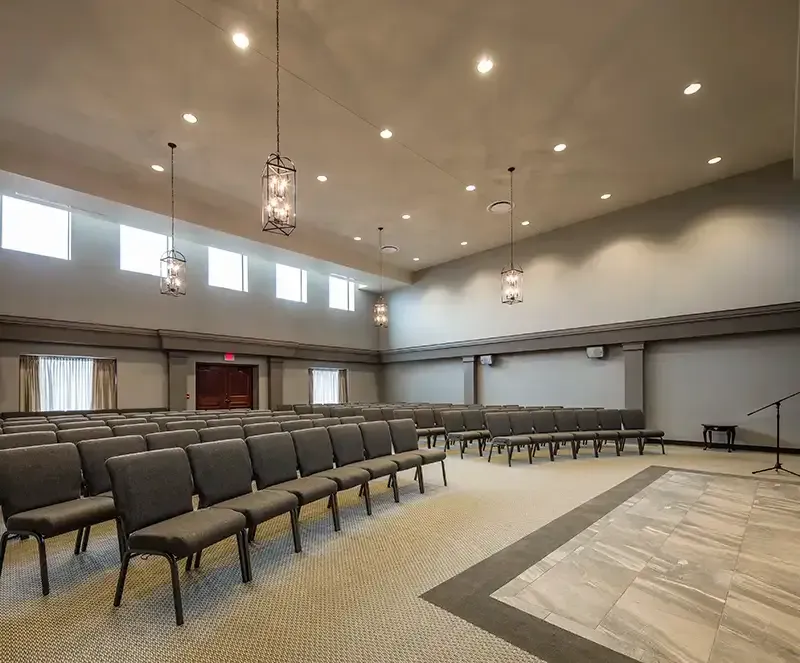 An empty, well-lit auditorium with rows of gray chairs. A stage with a microphone is on the right.