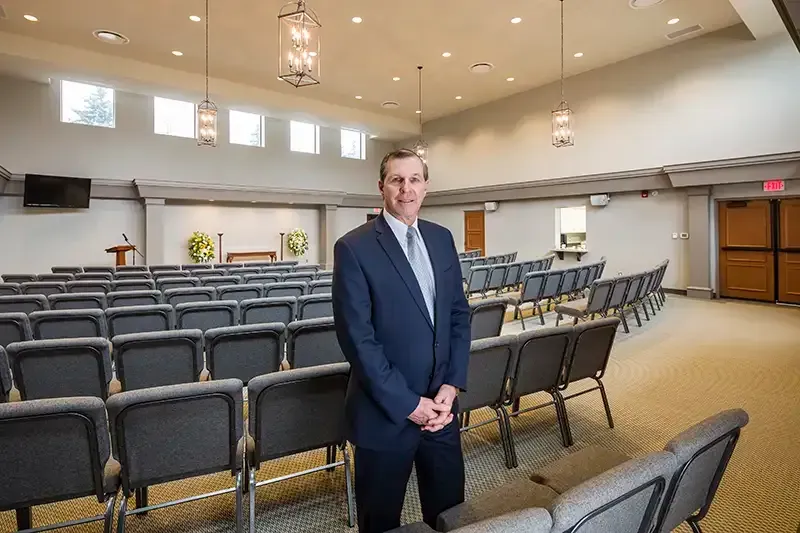 Man in suit stands in a large, empty funeral home chapel with rows of chairs.