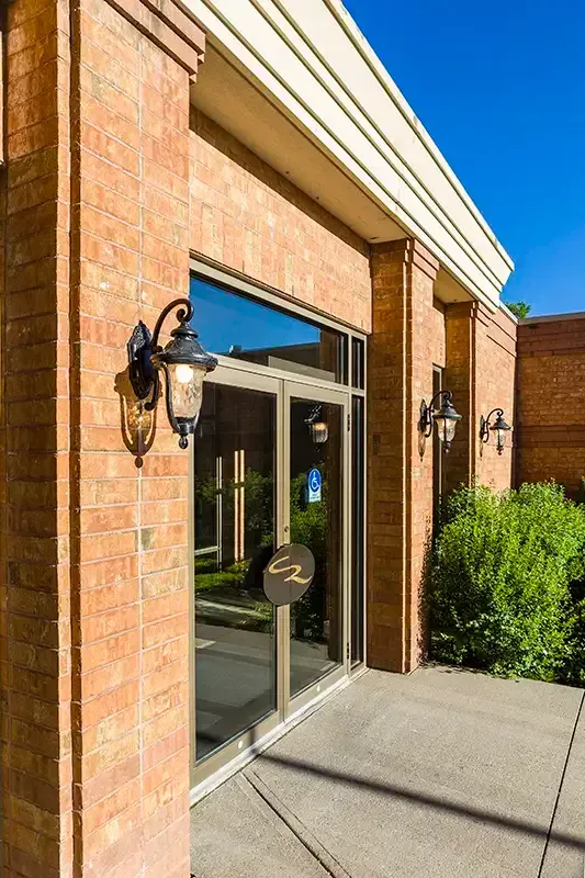 Entrance to a brick building with glass doors, sconces, and a small bush.