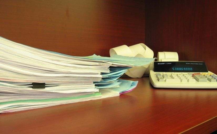 Stack of paperwork with a calculator and paper roll on a wooden desk.