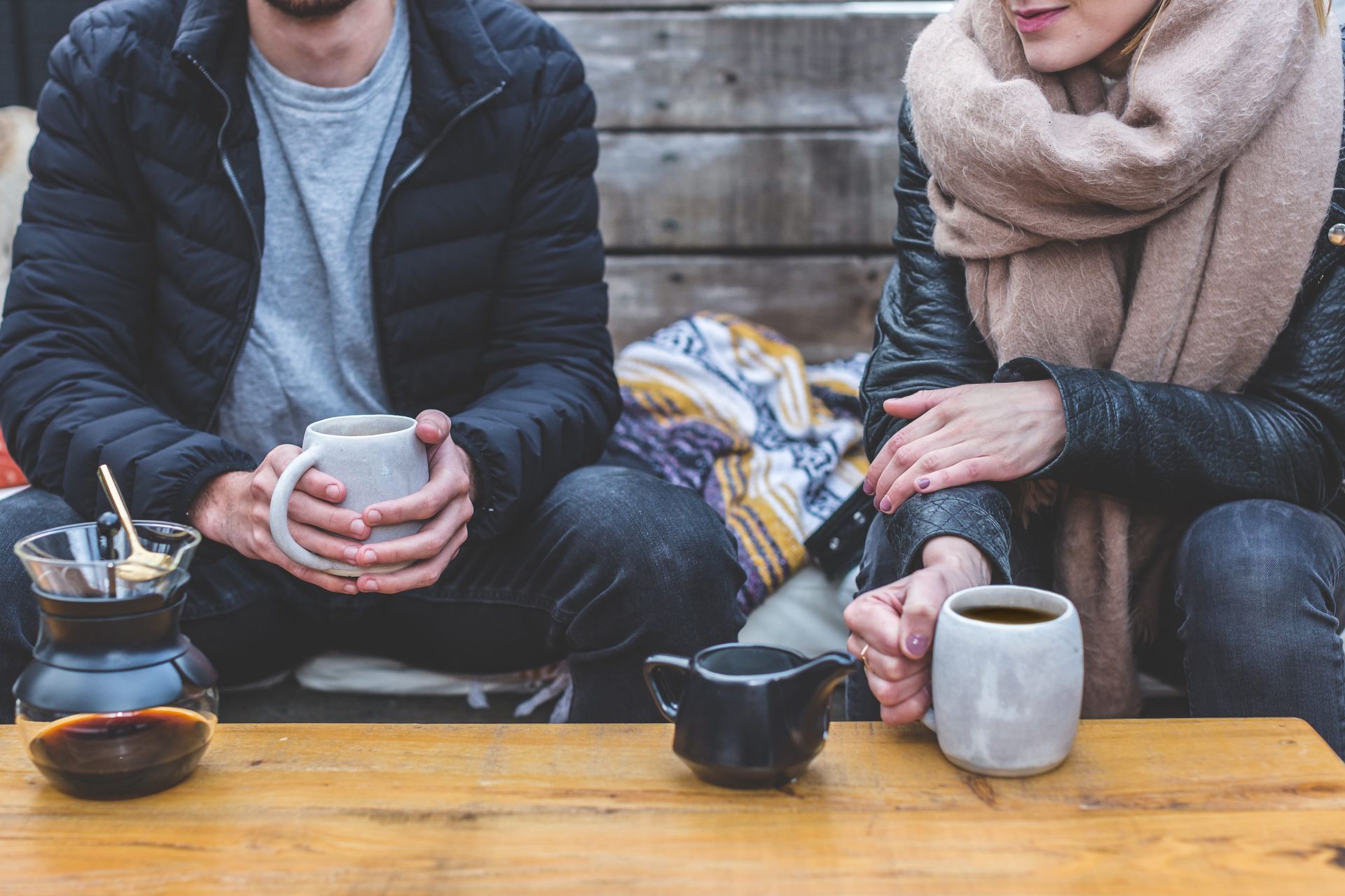 Two people, wrapped in coats and scarves, sit at a wooden table, drinking coffee outdoors.