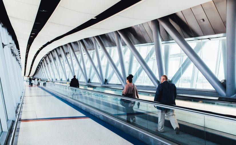 People walking on an airport moving walkway with a glass and steel exterior.