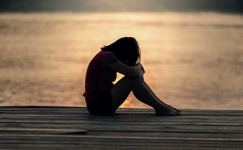 Woman, alone, sits on a wooden dock, head in hands, facing a sunset over water.