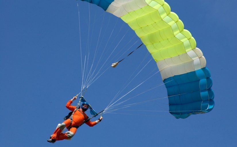 Skydiver in orange suit under a blue, white, and yellow parachute against a bright blue sky.