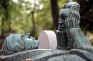 Bronze sculpture of two figures, one reclining, gazing at the other. Cemetery setting with trees.