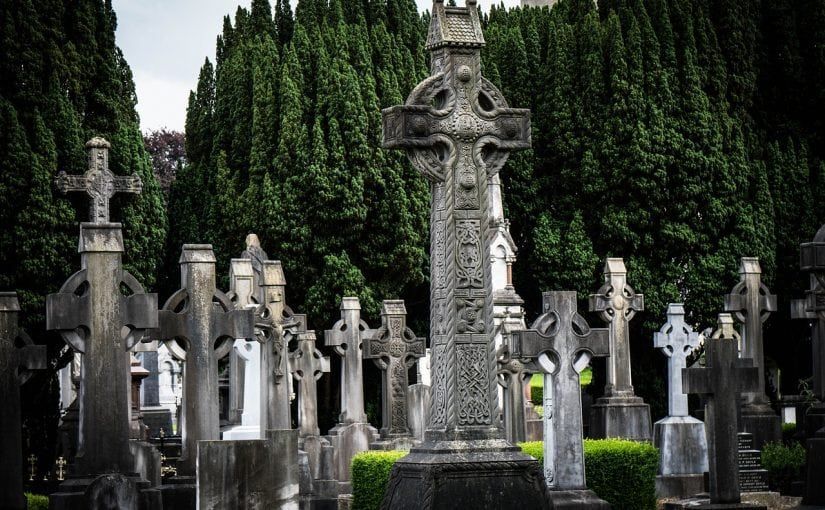 Celtic crosses in a graveyard, set against tall, dark green trees under a cloudy sky.