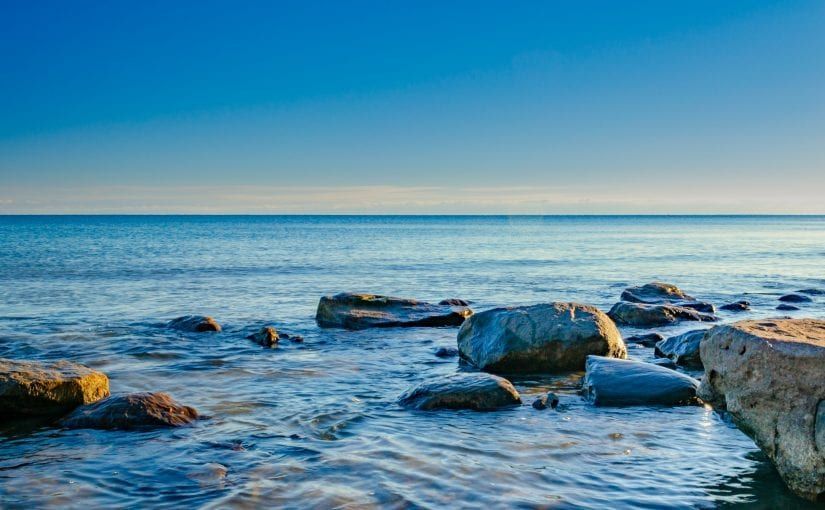 Blue ocean with rocks in the foreground, under a bright blue sky.
