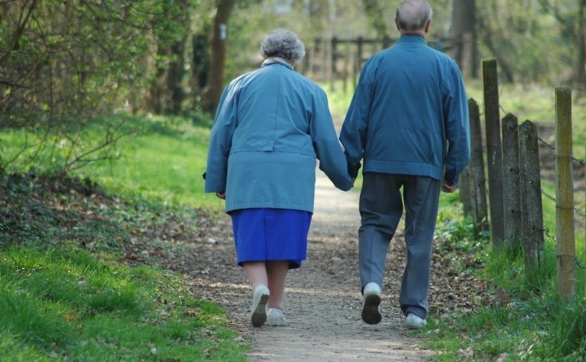 An elderly couple in blue jackets and skirt walk hand-in-hand on a path through a wooded area.