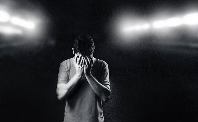Man with hands covering face, standing in darkness with bright lights in the background.