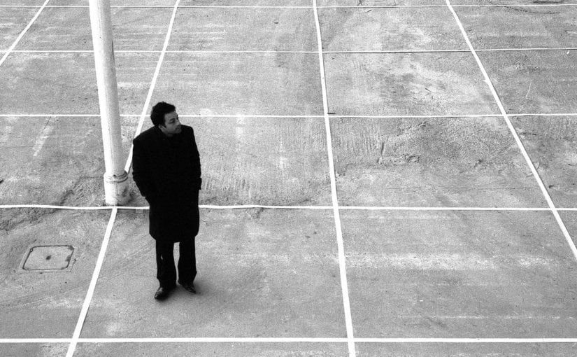 Man in black coat stands on a grid-marked concrete surface, near a white pole, looking off-camera.