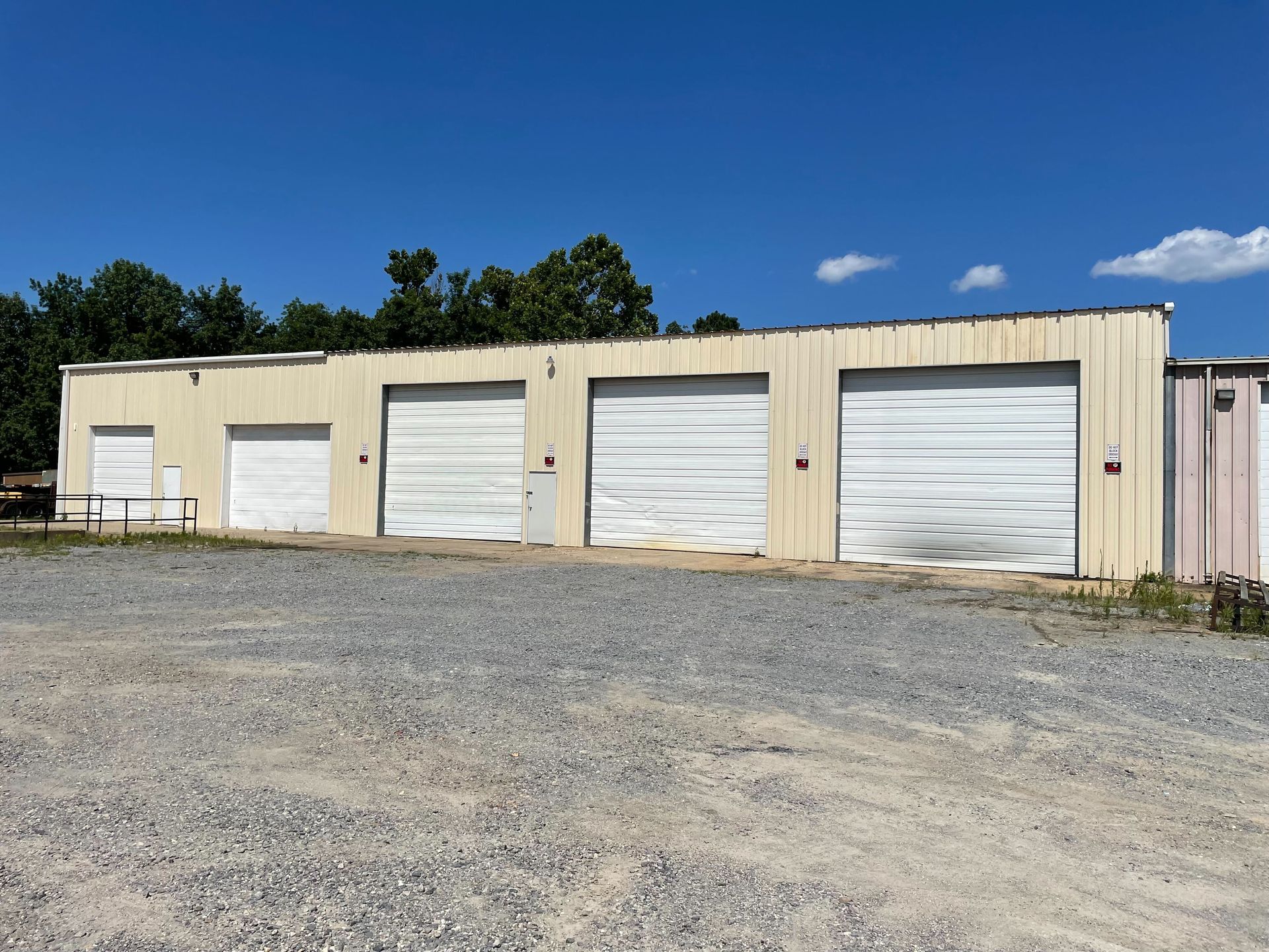 Light-colored commercial building with several garage doors under a blue sky, gravel lot in front.
