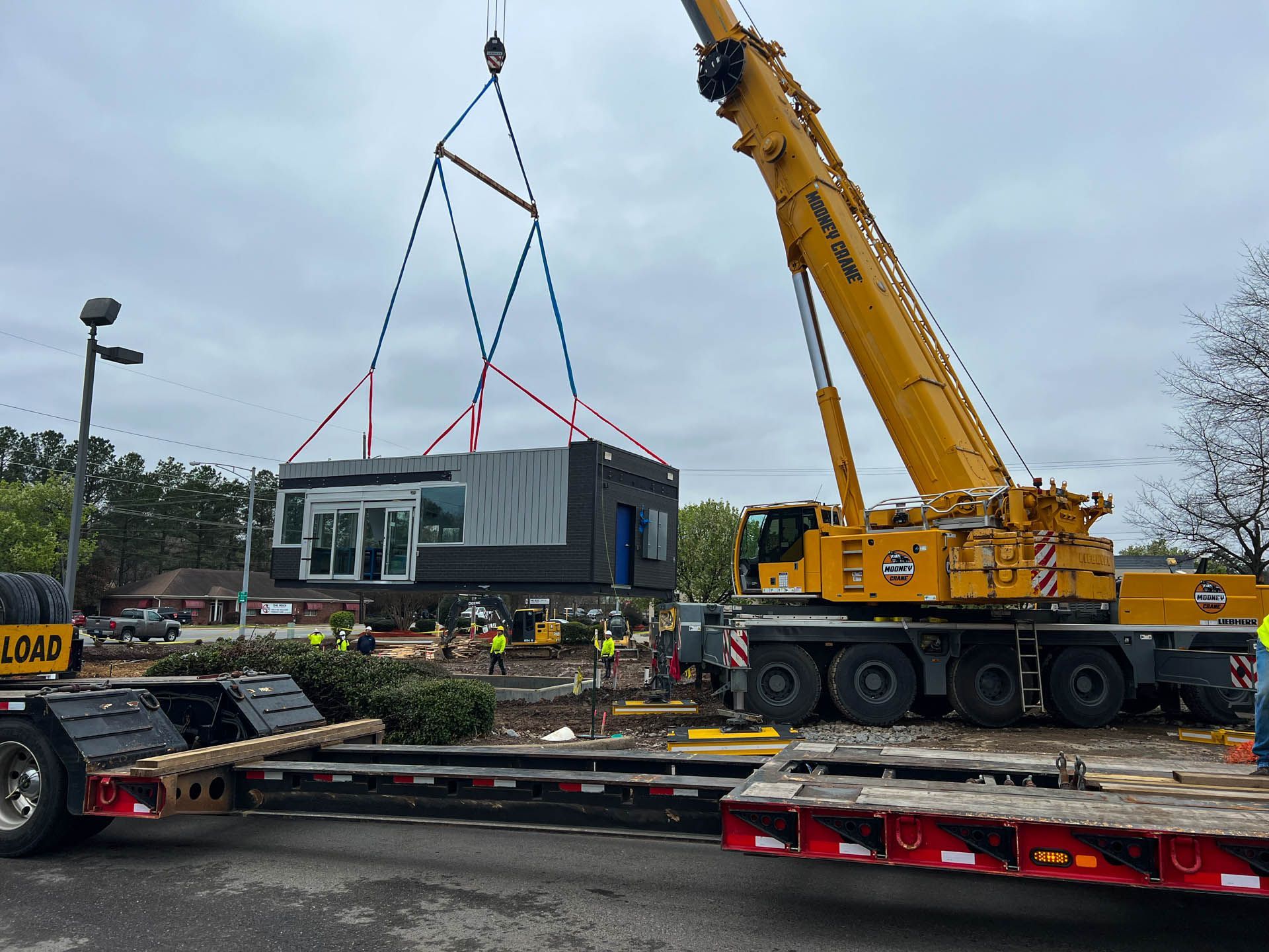 A large yellow crane lifting a modular building, workers on site, overcast day.