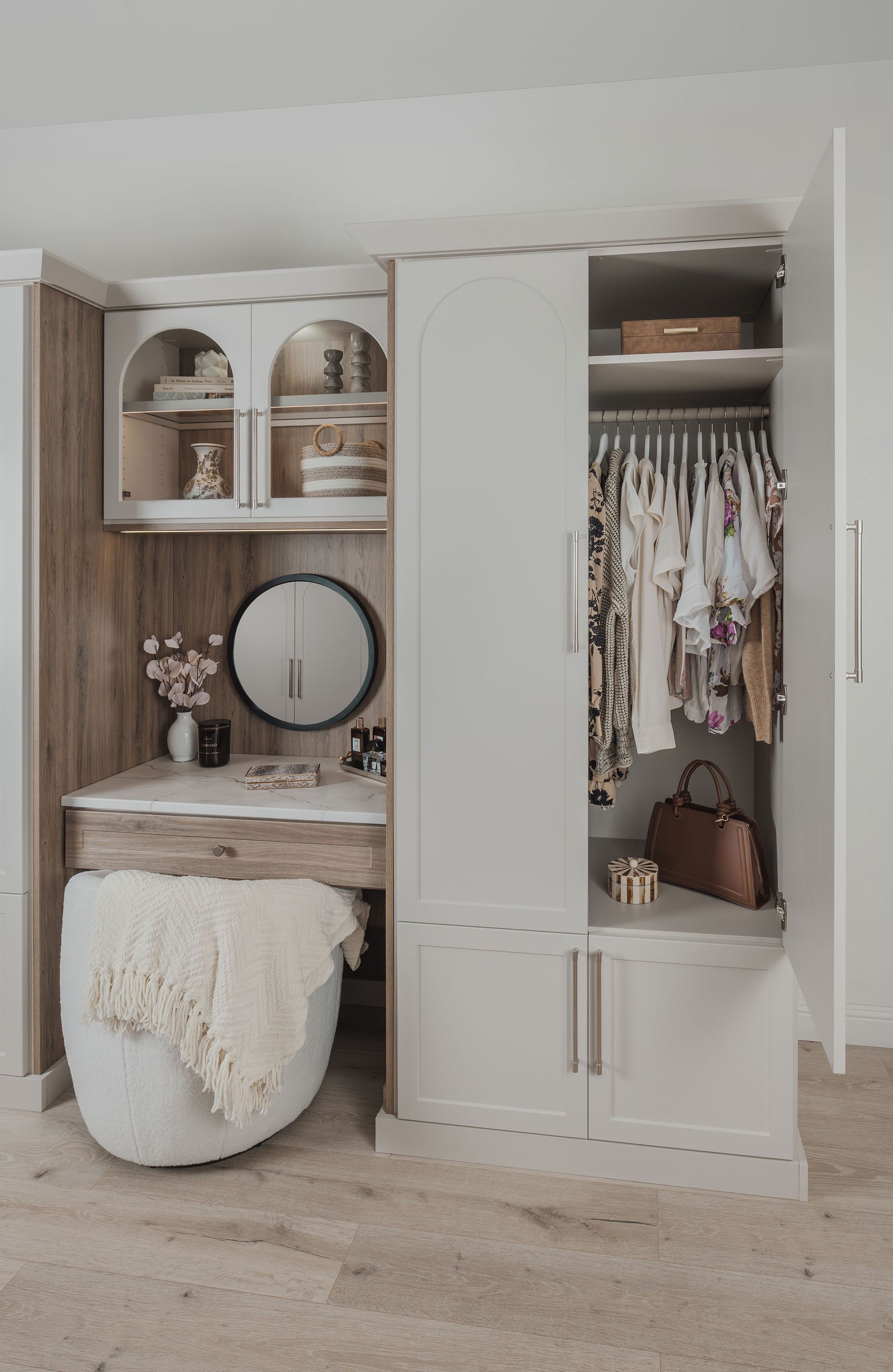 Built-in closet vanity with shelves, mirror, hanging clothes, and a white stool in a neutral bedroom.