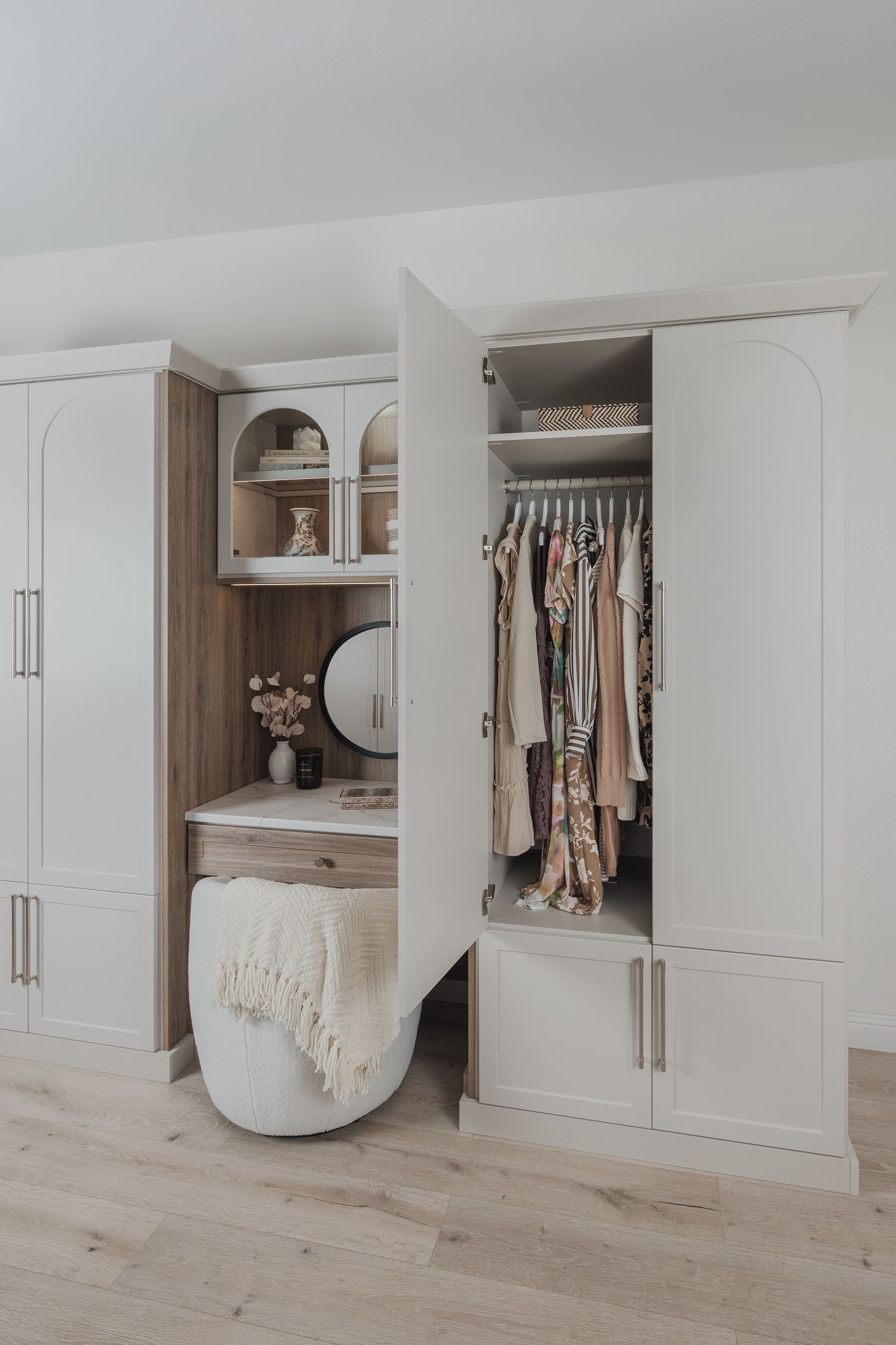 Built-in white closet with open doors, shelves, hanging clothes, and a vanity nook with a fluffy stool.