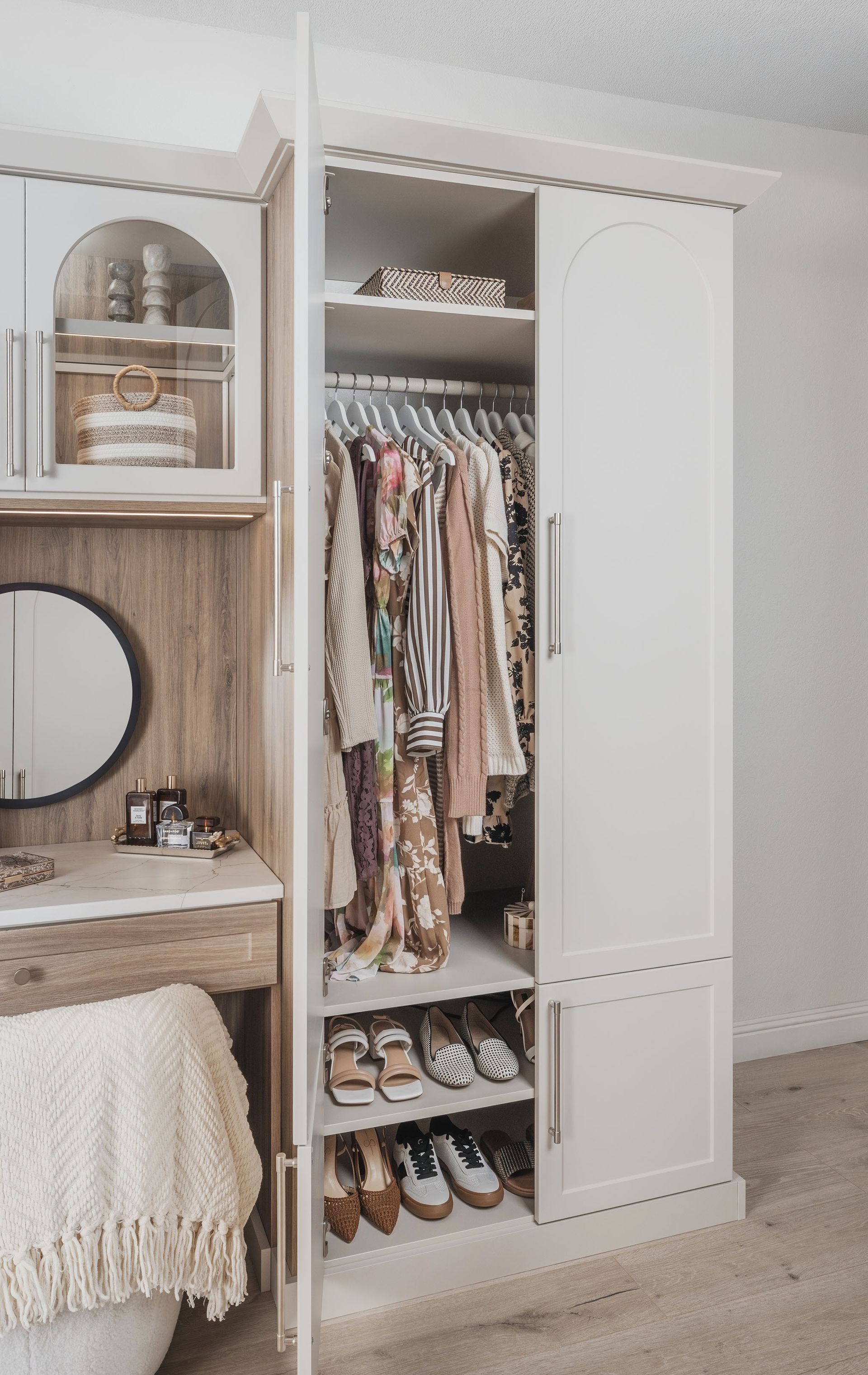 White wardrobe with open hanging clothes and shelves beside a wooden vanity in a bedroom