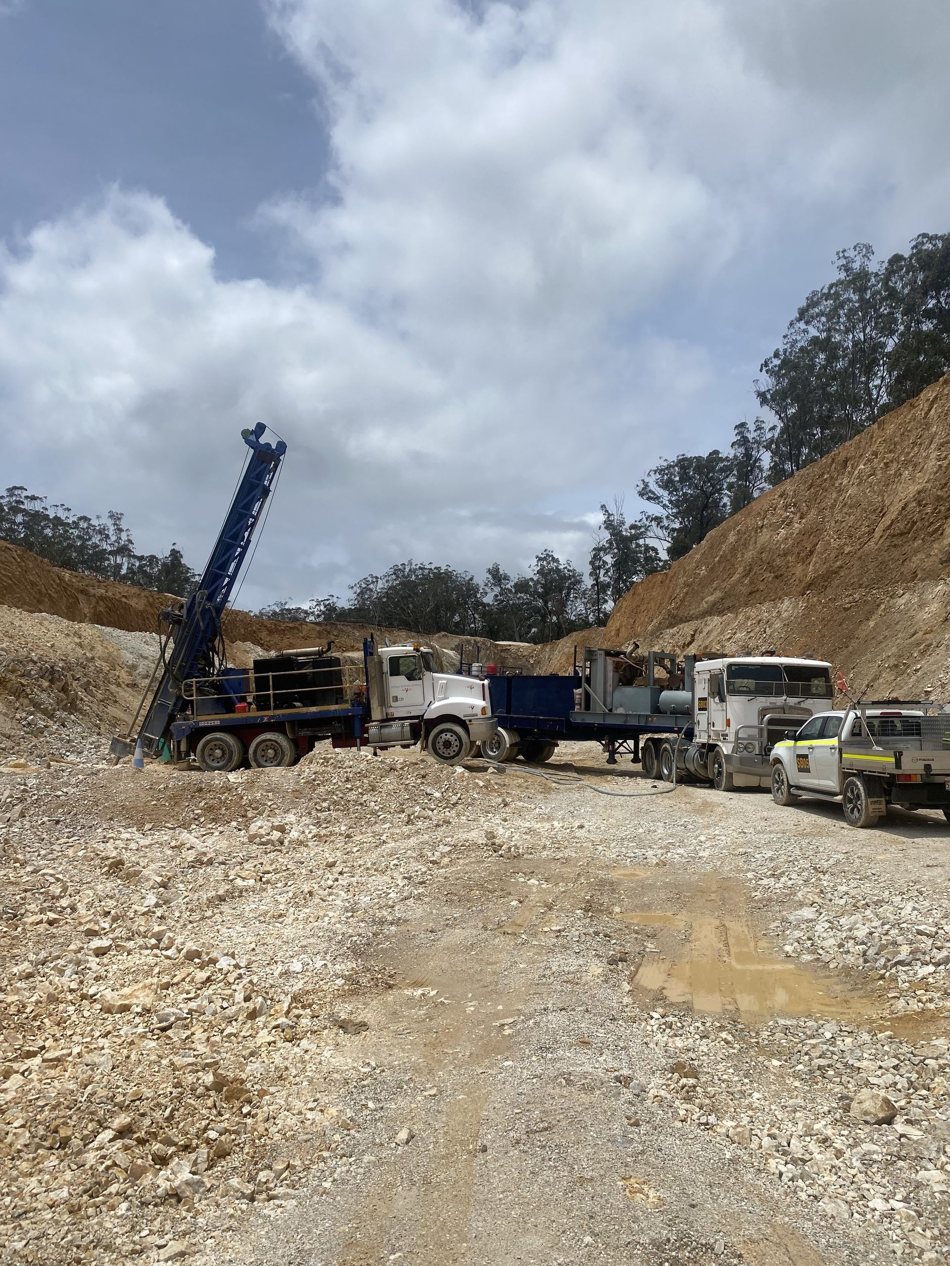 Drilling rig and trucks in a quarry, operating under a cloudy sky.