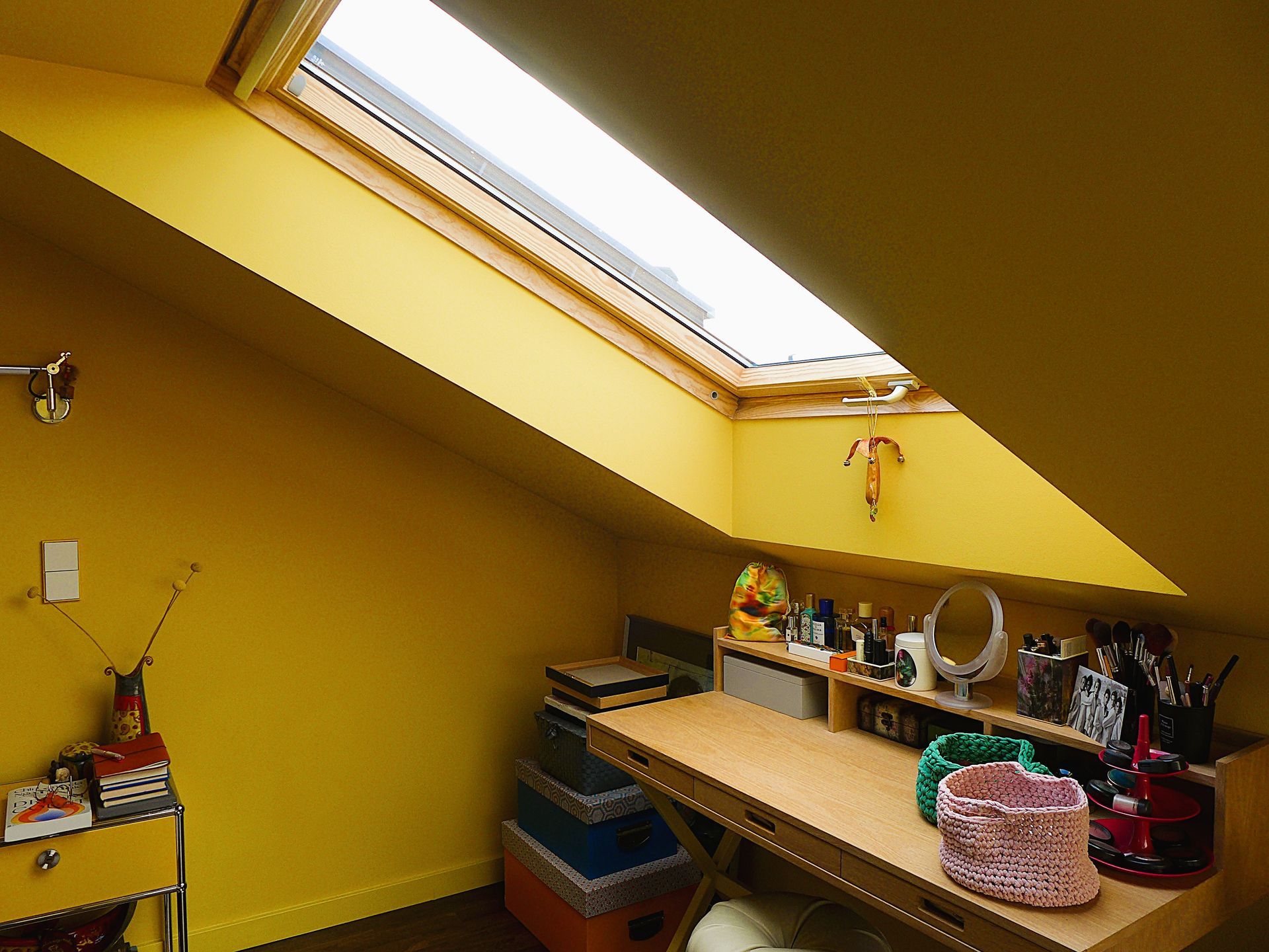 Ein Dachbodenbüro mit Dachschräge, Oberlicht und einem Schreibtisch, der mit Künstlerbedarf vollgestellt ist.