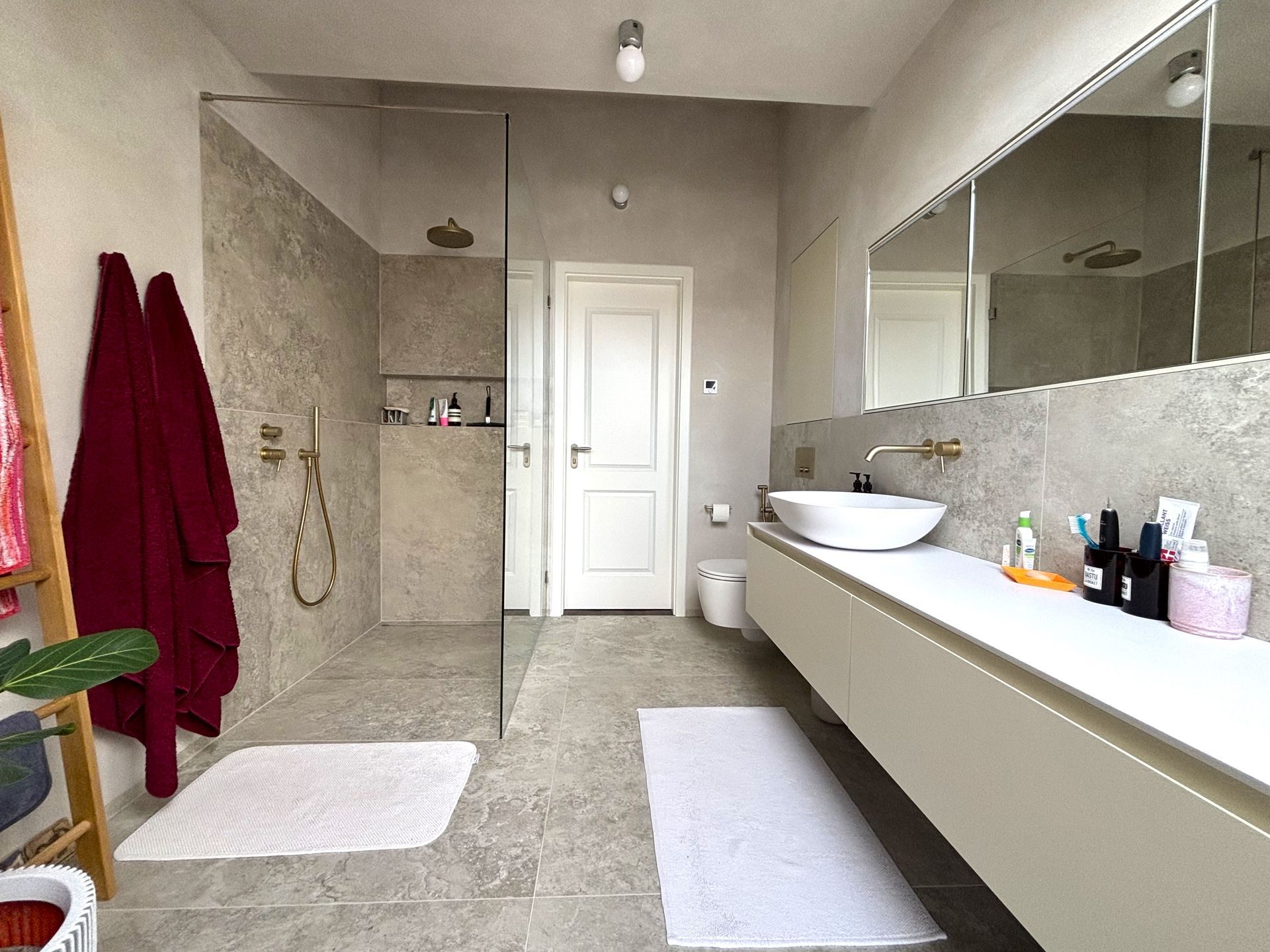 Modernes Badezimmer mit Dusche, langem, schwebendem Waschtisch mit Waschbecken und zwei weißen Teppichen auf dem Steinboden.