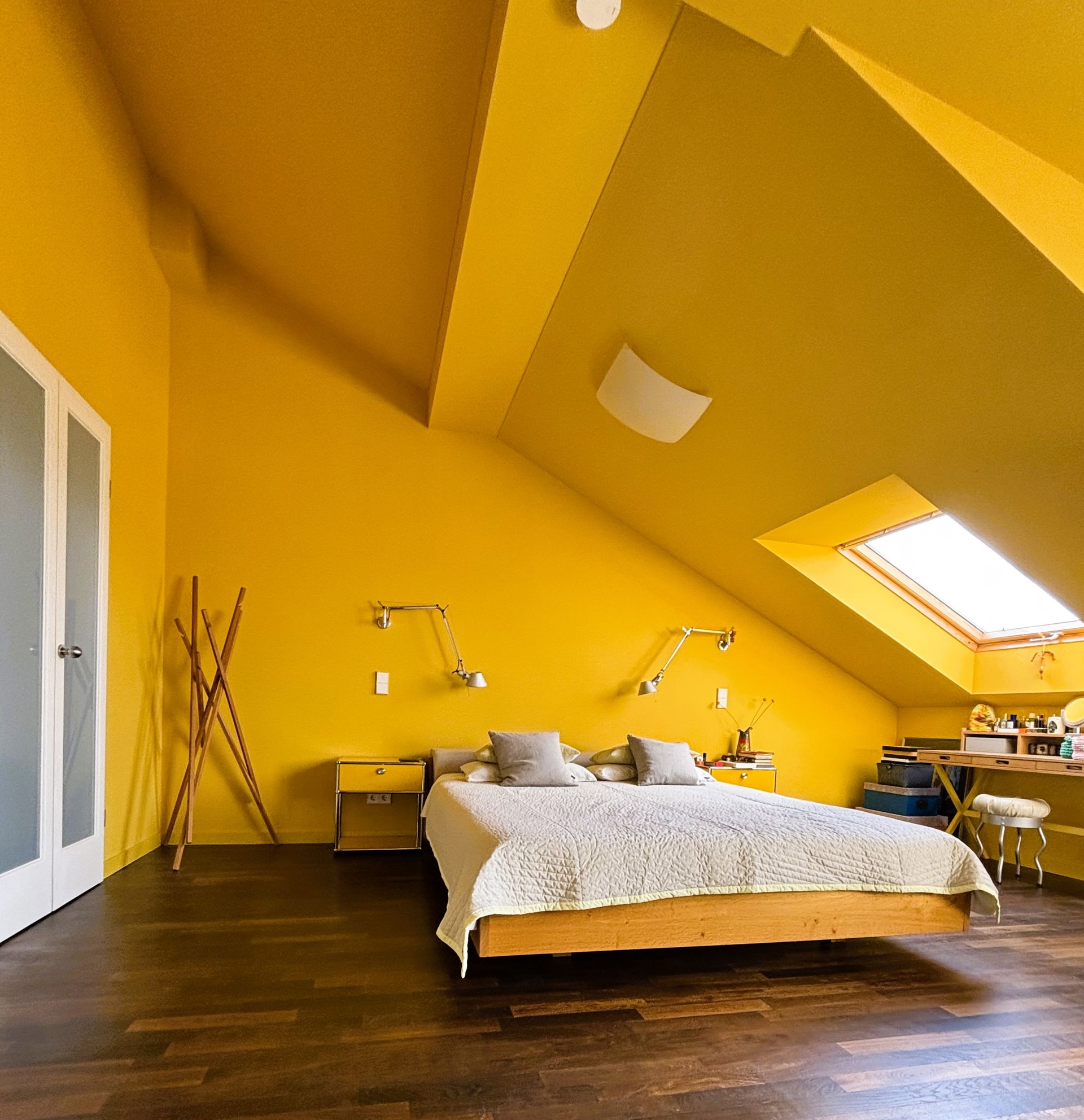 Ein Schlafzimmer mit leuchtend gelben Wänden, einem Holzbett, dunklen Dielen und einer Dachschräge mit Oberlicht.