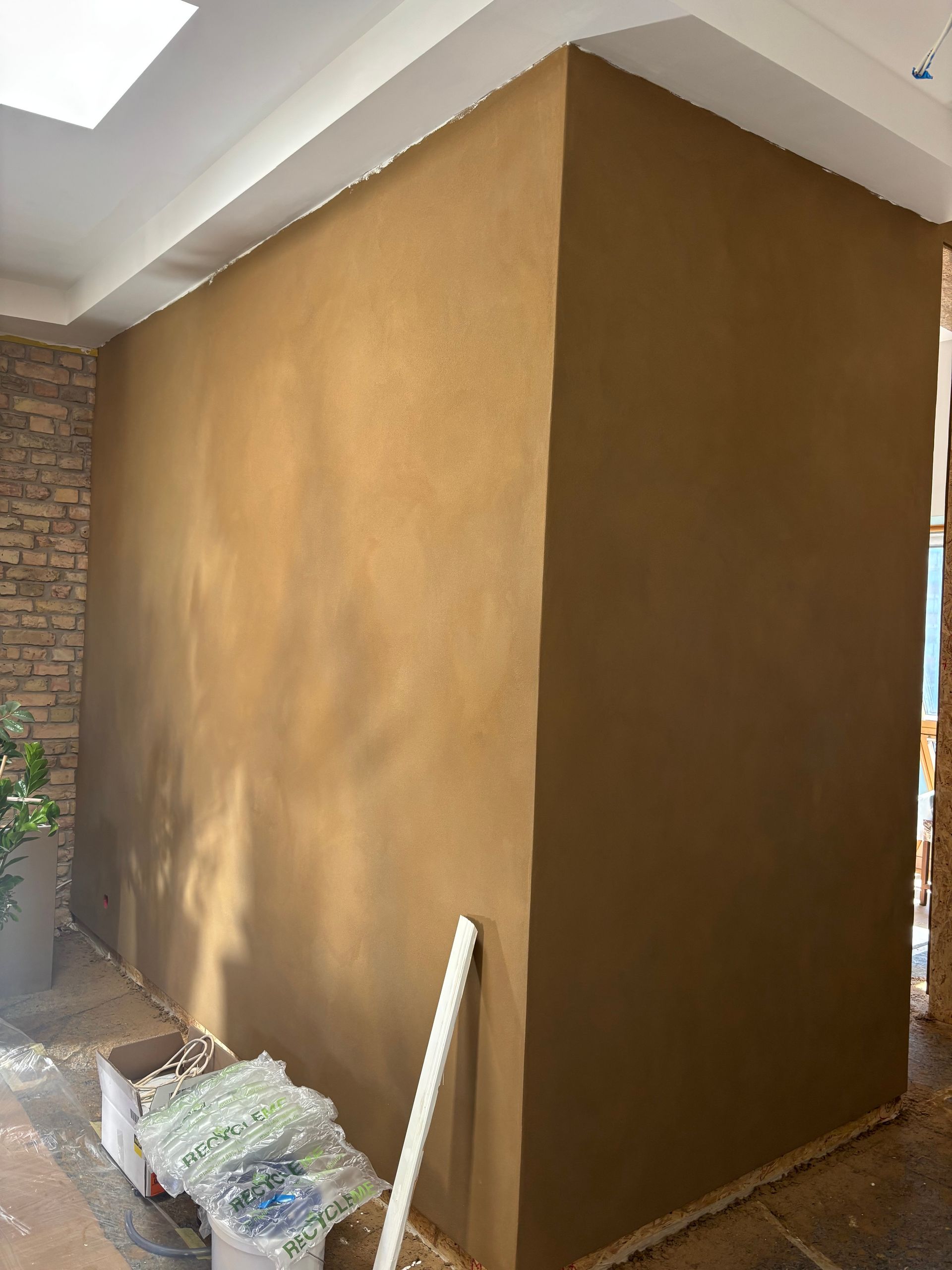 Eine frisch verputzte, glatte, hellbraune Wand bildet eine Ecke in einem Innenraum; daneben liegen Bauschutt auf dem Boden.
