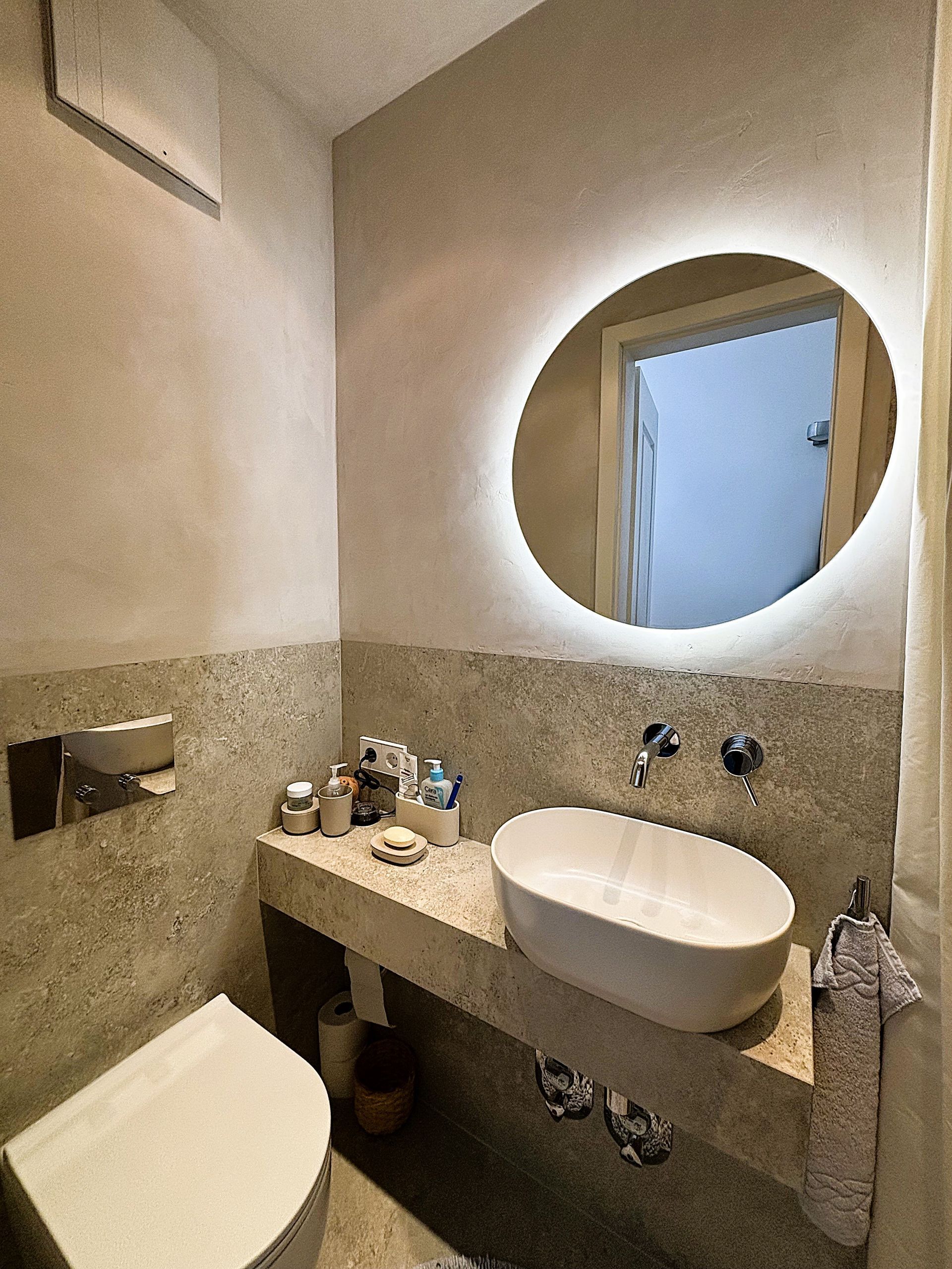 Ein Badezimmer mit einer Wand in Steinoptik, einem Aufsatzwaschbecken, einem runden Spiegel und einer Toilette.
