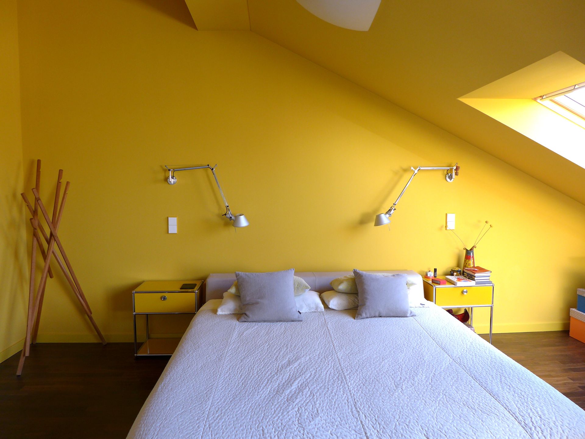 Ein hellgelbes Schlafzimmer mit Bett, zwei Nachttischen, Wandlampen und einer hölzernen Garderobe unter einer Dachschräge.