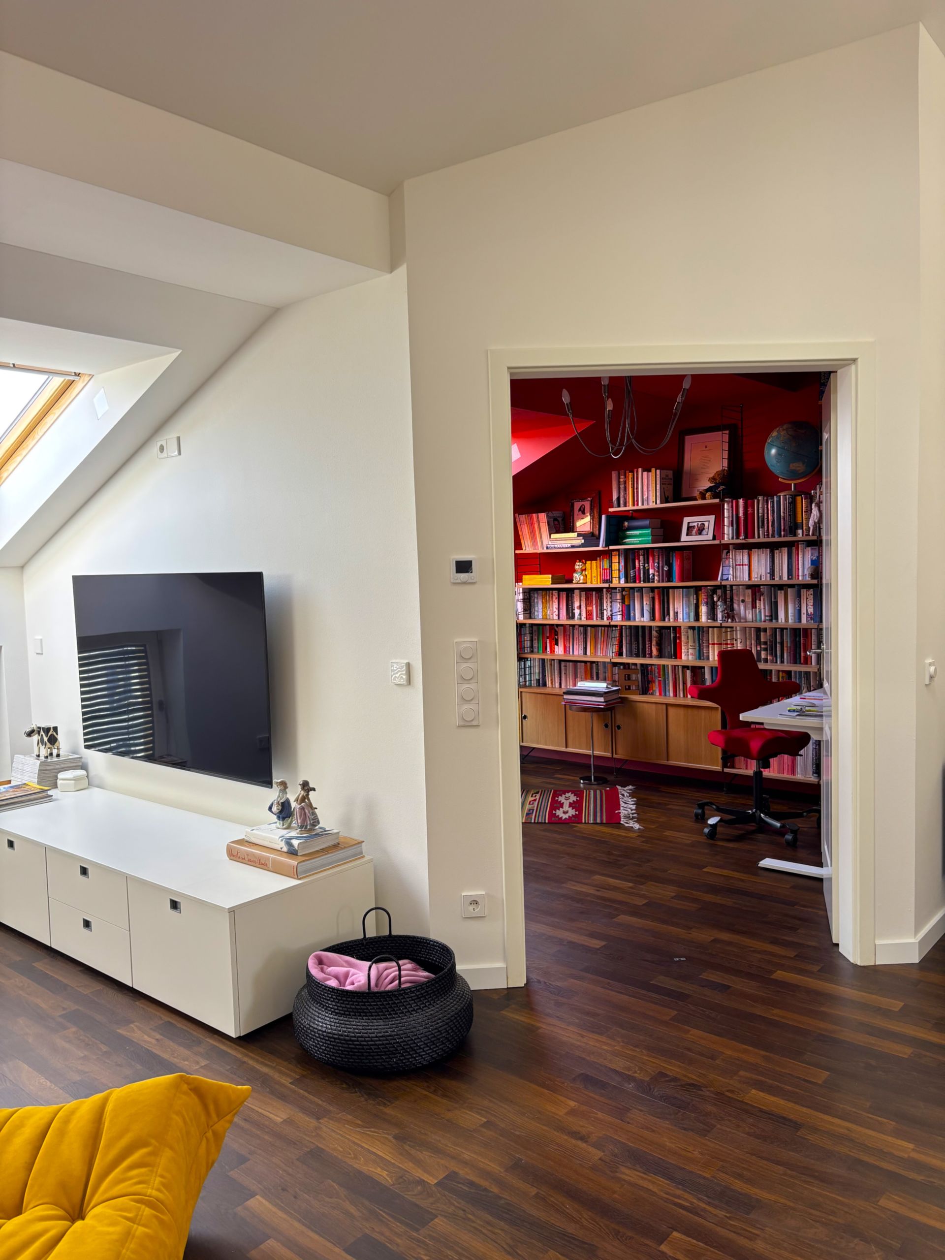 Ein Wohnzimmer mit einer weißen Medienkonsole, einem Fernseher und Blick in ein rotes Arbeitszimmer voller Bücherregale.
