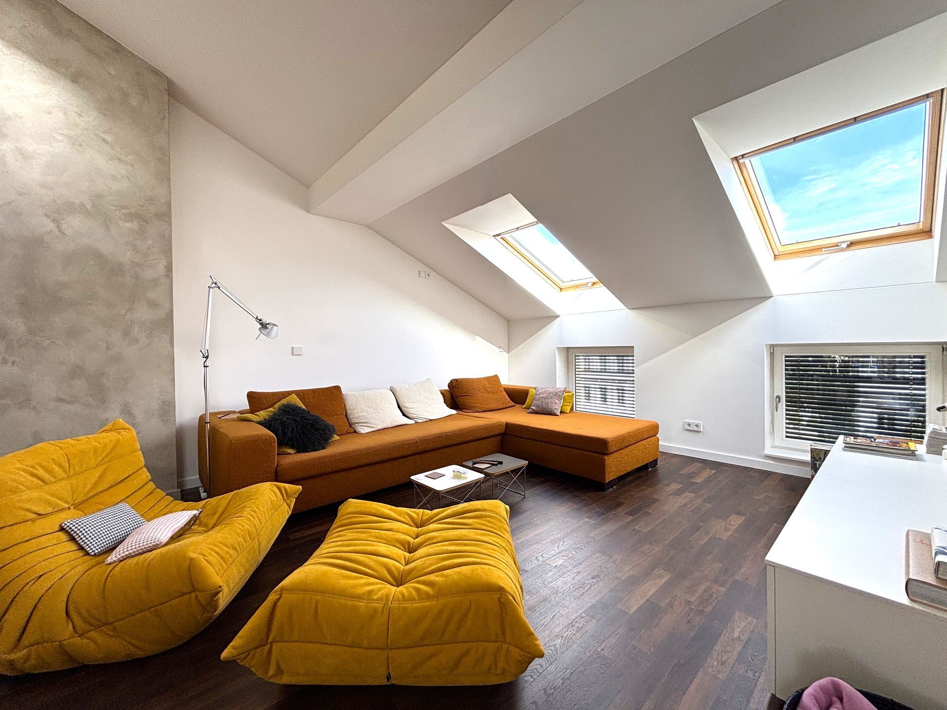 Ein loftartiges Wohnzimmer mit gelben Polstersesseln, einem braunen Ecksofa, Holzböden und mehreren Dachfenstern.