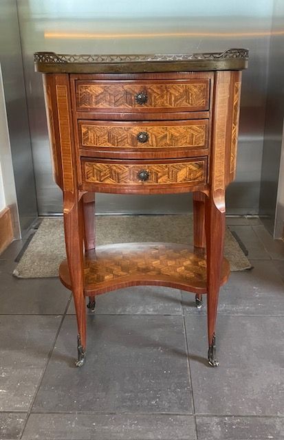 Ornate wooden side table with three drawers and curved legs against a metal wall.