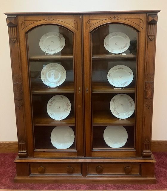 Wooden display cabinet with glass doors holding white plates on shelves and drawers below