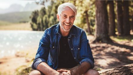 An older man is sitting on a rock near a lake and smiling.