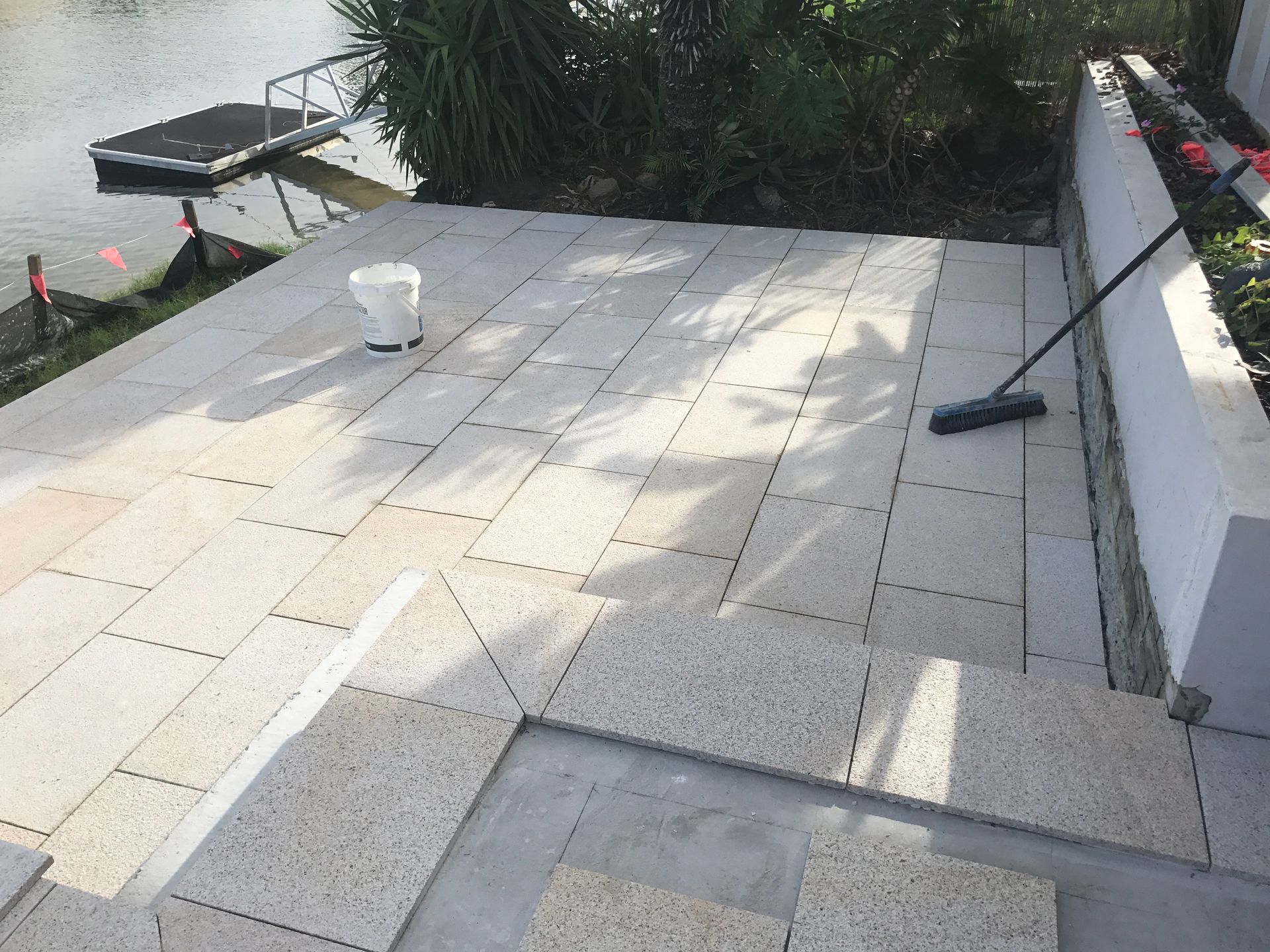 Light-coloured stone patio being installed beside a body of water. A bucket and broom sit on the patio. — Worrall Tiling in South Kempsey, NSW