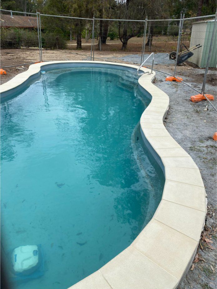 Swimming pool with blue water and beige stone coping, surrounded by a fence and dirt. — Worrall Tiling in South Kempsey, NSW