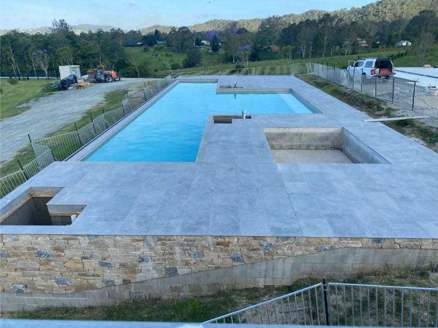 Rectangular pool under construction with stone surround; blue water, gray tiling, green grass, hills in background. — Worrall Tiling in South Kempsey, NSW