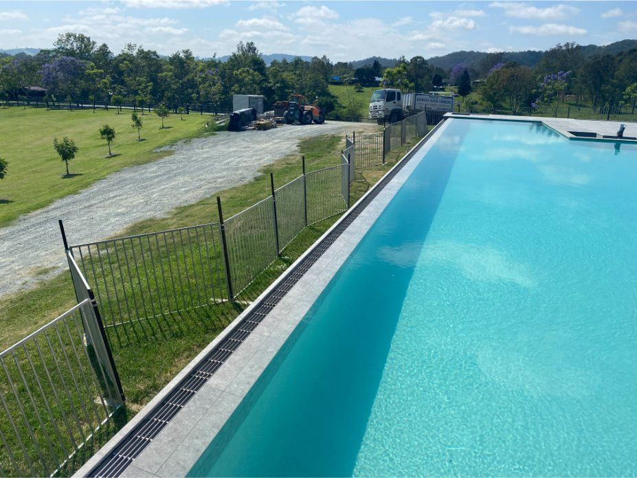 Long, turquoise swimming pool with black fence, gravel path, trees, and blue sky. — Worrall Tiling in South Kempsey, NSW