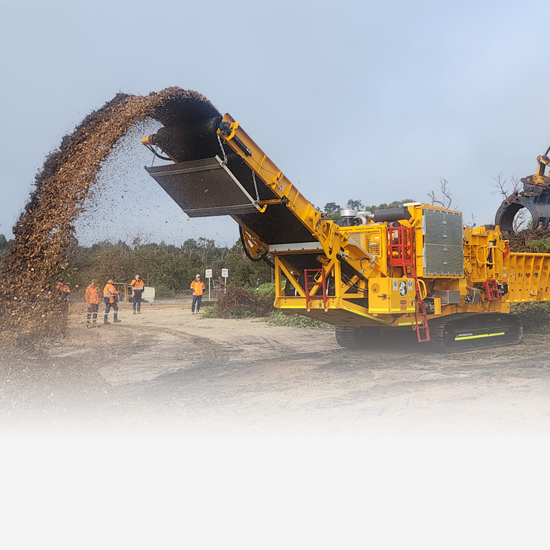 Yellow industrial wood chipper unloading a large arc of wood chips at a worksite with workers nearby