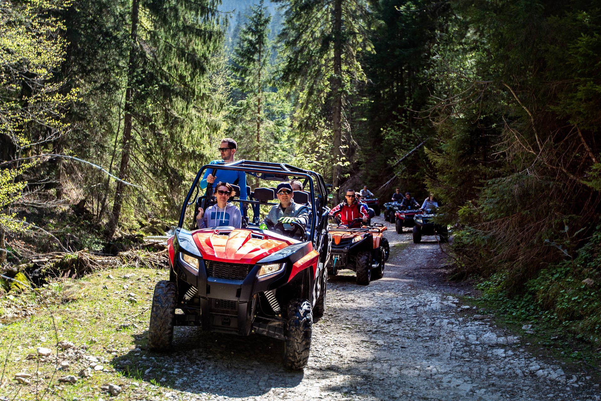 ATVs driving on a forest trail, with a red lead vehicle and riders in helmets.